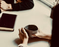 Person holding a cup at a table with another person and a notebook in the background.