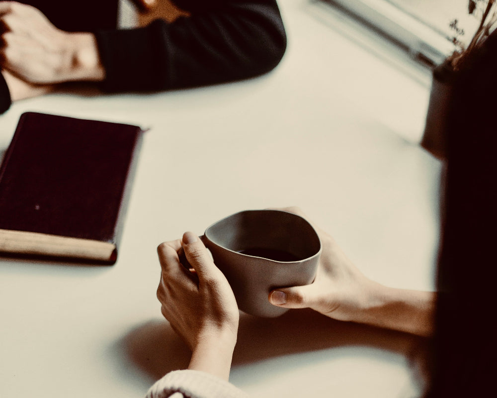 Person holding a cup at a table with another person and a notebook in the background.