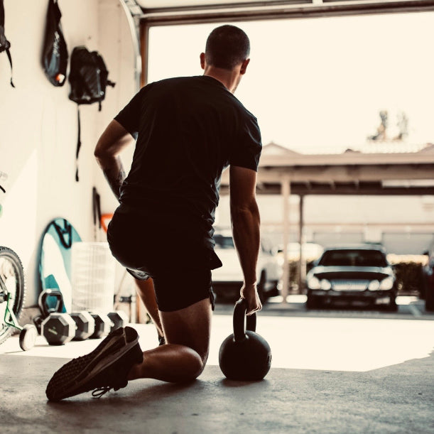 A man in a black t-shirt and shorts kneels on a garage floor holding a kettlebell, viewed from behind as he faces an open garage door with cars outside.